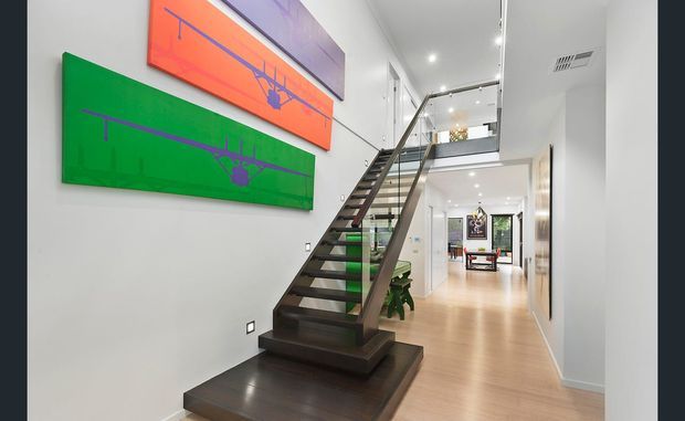 Bright contemporary interior hallway featuring a sculptural dark timber open-riser staircase with glass balustrade leading to an upper landing. White walls, pale timber flooring, recessed lighting, and vivid green, orange, and purple abstract artwork create a colourful modern gallery-like setting.