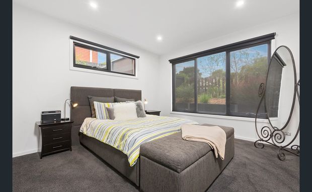 A bright, modern bedroom with a dark upholstered bed, matching footstool, grey carpet, white walls, black-framed windows, and layered neutral bedding with subtle yellow and blue stripes. A black bedside table and decorative full-length mirror complete the room.