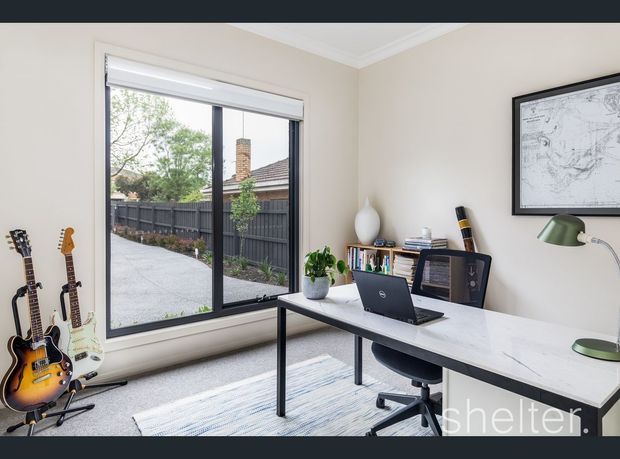 Bright, compact home office with a modern white desk, laptop, ergonomic office chair, green desk lamp, books, and potted plant. A large black-framed window provides views of a fenced outdoor area, while several guitars are displayed on stands along the wall. Neutral walls, carpeted flooring, and framed artwork create a clean, contemporary workspace.