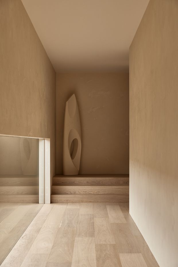 A narrow, minimalist interior passage with warm beige plaster walls and ceiling, pale timber flooring, and a short set of timber steps leading to a recessed alcove. A tall sculptural object stands in the niche, while a large mirror or reflective panel on the left extends the view. The space is softly lit and monochromatic, with clean architectural lines and built-in detailing.