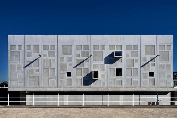 A frontal exterior view of a large, rectilinear modern building under a deep blue sky. The facade is composed of pale grey modular concrete panels with a dense geometric grid of recessed and projecting rectangular window openings, some casting strong angular shadows. A darker lower level is screened by horizontal metal railings, and the building sits on a paved urban forecourt.