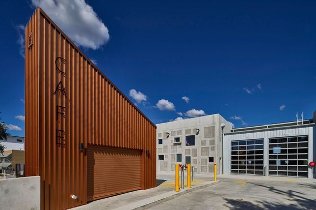 Exterior architectural view of a contemporary industrial or commercial complex under a vivid blue sky. The foreground building has a tall, narrow form clad in warm rust-orange vertical corrugated metal, with a large roller door and subtle vertical signage. Adjacent low-rise buildings use pale concrete and white metal façades, with extensive glazed sectional garage or workshop doors, service bollards, and a paved forecourt.