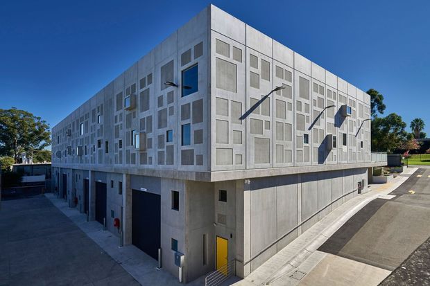 Exterior view of a large contemporary multi-storey concrete building with a strongly geometric facade. The upper levels feature a grid of recessed and projecting rectangular panels, windows, and external sun-shading elements, while the lower level appears to contain service or parking access. The building sits beside a paved driveway and streetscape under clear blue skies.