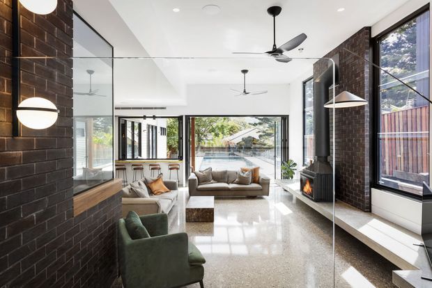 Bright contemporary open-plan living room with polished terrazzo flooring, dark exposed brick feature walls, extensive glazing, a large mirrored wall, ceiling fans, pendant lighting, a fireplace, and a compact bar or kitchen area at the rear. The room opens visually toward a pool and landscaped outdoor area.