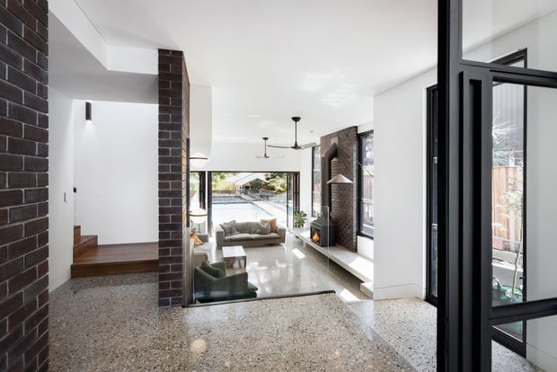 Bright contemporary open-plan living area viewed from an entry or hallway, featuring polished terrazzo flooring, exposed dark brick columns and fireplace wall, white walls, black-framed glazing and doors, timber steps, and a sunken lounge with a sofa and armchairs. The room opens toward a landscaped outdoor area through large windows.