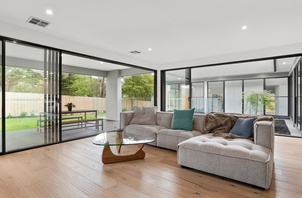 Bright contemporary living room with expansive black-framed floor-to-ceiling glazing, pale timber flooring, white walls and ceiling, and a large L-shaped sectional sofa arranged around a sculptural wood-and-glass coffee table. The glazing opens visually onto a landscaped courtyard and covered outdoor area.