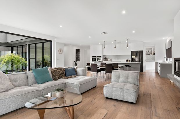 Bright open-plan contemporary interior combining a living room, dining area and kitchen. The space features light timber flooring, white walls and cabinetry, black-framed floor-to-ceiling glazing, a large pale sectional sofa, glass coffee table, dining table, and a central kitchen island with pendant lights. A fireplace and built-in shelving are visible at the right.