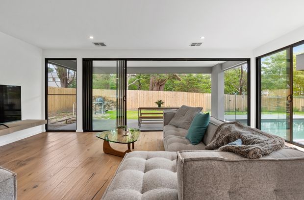 Contemporary living room with a large grey sectional sofa, glass coffee table, timber flooring, and extensive black-framed sliding glass doors opening to a landscaped backyard and pool. The room is bright and connected to the outdoor area, with a clean neutral palette and abundant natural light.