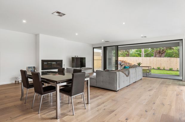 Bright open-plan living and dining area with pale timber flooring, white walls and ceiling, a dark dining table with upholstered chairs, and a grey sectional sofa facing a fireplace and television. Large black-framed sliding glass doors open to a fenced backyard and patio, creating a strong indoor-outdoor connection.