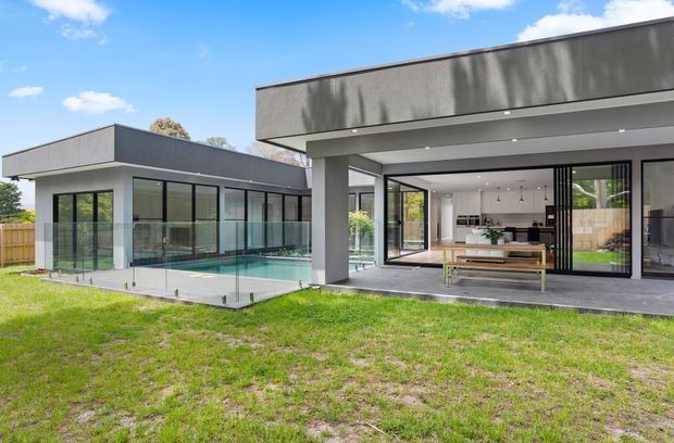 Contemporary single-storey residence with a flat-roofed, dark-rendered exterior and extensive black-framed glazing. The view looks across a lawn toward a glazed pool courtyard and an open-plan living and kitchen area, with a covered outdoor entertaining zone and frameless glass pool fencing.