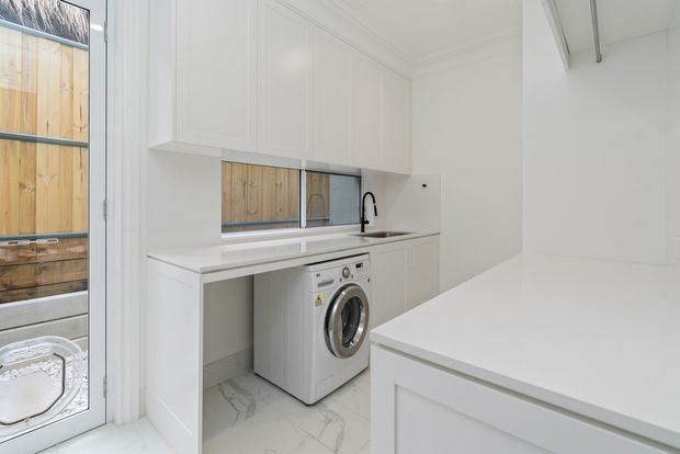 Bright, compact white laundry room with built-in cabinetry, a long benchtop, integrated sink and black tap, front-loading washing machine, and marble-look floor tiles. A large white island or folding bench occupies the foreground, with timber screening visible through the glazing.