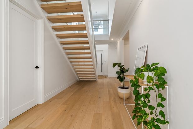 Bright, minimalist hallway with a floating timber staircase, white painted walls and panelled doors, light timber flooring, and a skylit void above. Indoor plants and a leaning mirror add natural accents to the clean contemporary space.