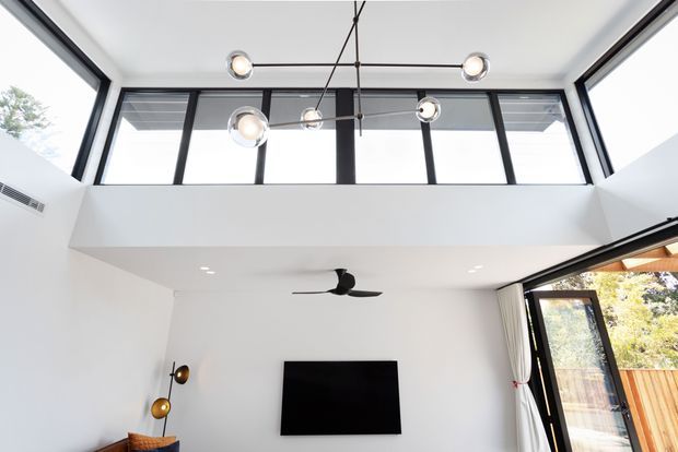 Bright contemporary living room with a double-height ceiling, clerestory glazing, and large black-framed windows and doors. A black ceiling fan, wall-mounted television, and sculptural multi-light pendant occupy the predominantly white space, with timber fencing and greenery visible outside.
