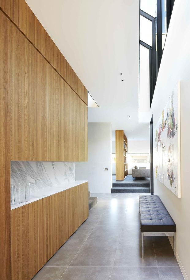 A bright, narrow contemporary interior passage with extensive vertical timber cabinetry and wall panelling on the left, a white marble-look splashback, large grey floor tiles, and a long tufted upholstered bench along the right wall. Black-framed double-height glazing and a skylit void bring in natural light, with a living area visible at the far end. A large abstract artwork hangs on the right wall.