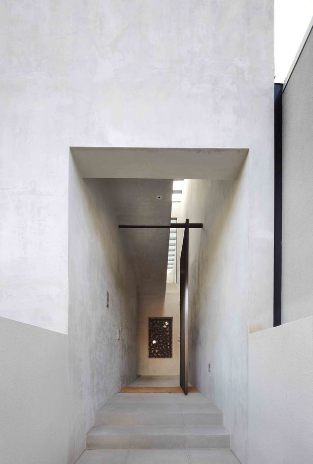 A minimalist, narrow interior passage reached by a short flight of pale steps. White textured plaster walls and ceiling form a deep recessed corridor, with a linear skylight bringing daylight from above. A dark slim structural or door element crosses the passage, and a decorative perforated panel is visible at the far end. The space has a stark, calm, gallery-like character with strong vertical and horizontal geometry.