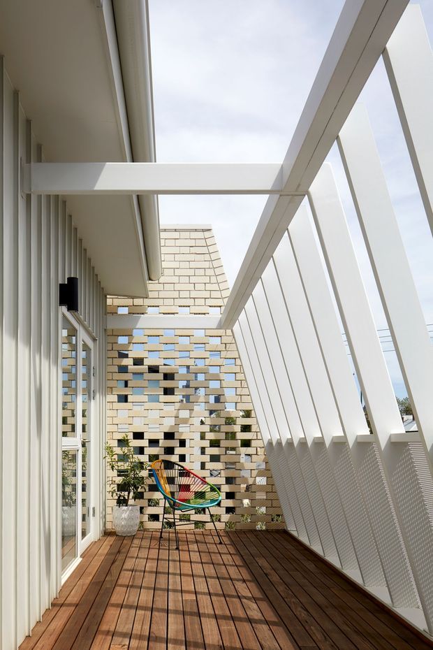 A bright, narrow elevated balcony or terrace with warm timber decking, framed by white structural beams and a strong angled white balustrade with perforated panels. Full-height glazed doors and vertical screening line one side, while a perforated cream brick privacy wall encloses the far end. A colourful outdoor chair and potted plant provide small accents. The space has a clean contemporary architectural character with strong geometric lines and filtered daylight.