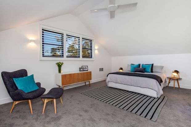 Bright attic-style bedroom with a sloping white ceiling, grey carpet, and a large bed dressed in white and grey bedding. The room includes teal accent cushions, a dark upholstered lounge chair and ottoman, a timber sideboard, striped rug, bedside table, wall lights, and a ceiling fan. Plantation shutters frame a window overlooking trees.