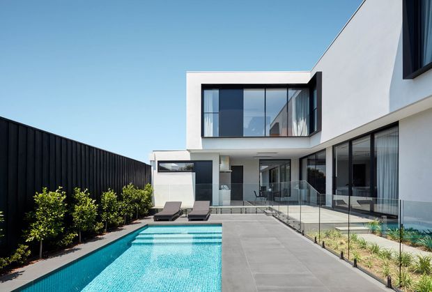 Contemporary two-storey residence with crisp white rendered walls, extensive black-framed glazing, and a rectilinear flat-roof form. The image looks across a paved courtyard toward the house, with a long narrow swimming pool, glass balustrade, dark boundary fencing, and integrated outdoor seating. The composition is bright and minimal, emphasizing strong geometric lines and indoor-outdoor connection.