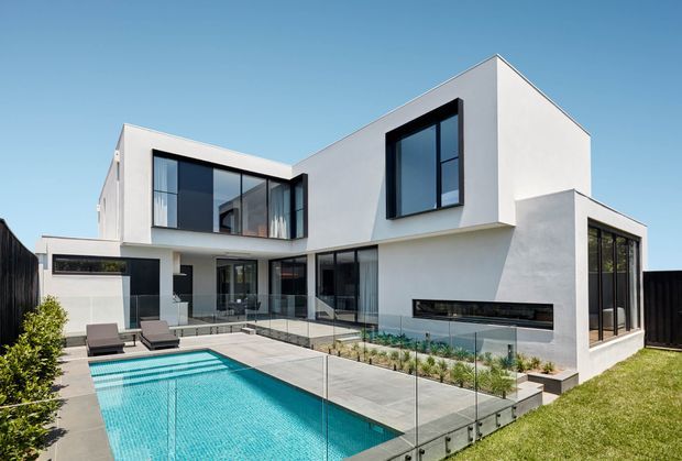 Contemporary two-storey white-rendered home with strong cubic forms, expansive dark-framed glazing, a recessed outdoor entertaining area, linear built-in fireplace, landscaped courtyard, and rectangular swimming pool. The image is photographed from the rear garden/pool area under clear daylight.