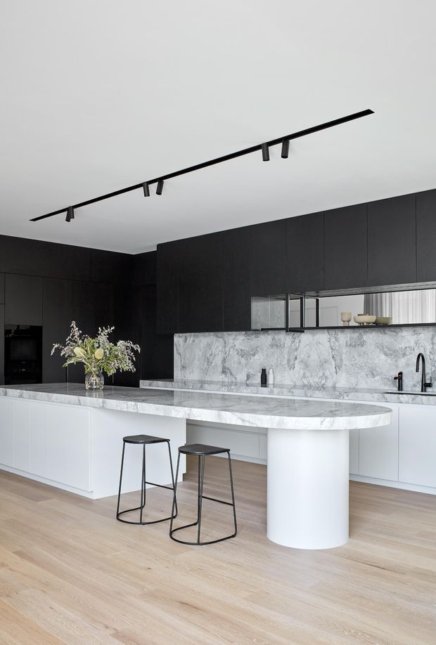 A refined contemporary kitchen with a large white marble island and rounded cylindrical support, pale timber flooring, white lower cabinetry, and extensive matte black upper cabinetry. A long linear black track light runs across the white ceiling, while a marble splashback, black tapware, minimalist stools, and a mirrored display niche add contrast. The space is bright, clean, and highly considered.