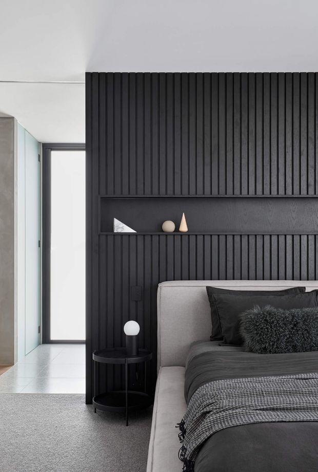Contemporary bedroom with a strong monochromatic palette. A grey upholstered bed sits against a dramatic black vertical timber-slat feature wall with an integrated recessed display shelf. Black bedding, a textured dark cushion, round black bedside table, and spherical lamp create a refined, minimalist look. A bright hallway or dressing area is visible to the left.