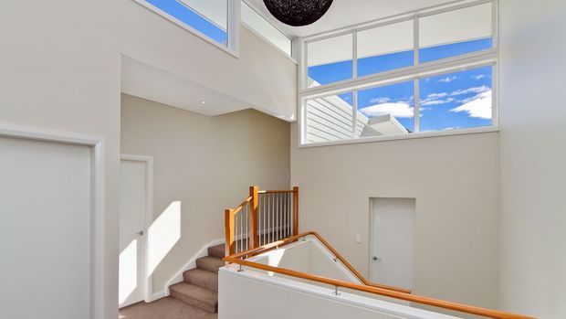 Bright modern stairway and upper landing with a double-height void, pale walls, carpeted stairs, and warm timber handrails with metal balustrades. Large high-level windows and clerestory glazing flood the space with natural light and reveal blue sky beyond.