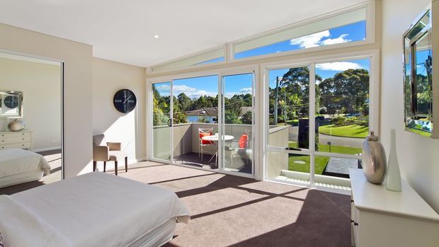 Bright contemporary bedroom with a large white bed, neutral carpet, white walls, and extensive floor-to-ceiling glazing opening onto a balcony. The glazing provides views of a leafy suburban landscape, while a simple console, ceramic vases, wall clock, and armchair complete the calm interior.