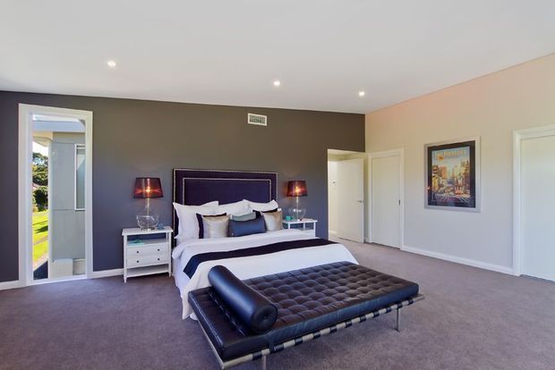 A spacious, softly lit bedroom with a large upholstered bed centered against a dark feature wall, matching bedside tables and lamps, and a tufted leather bench at the foot of the bed. The room has pale painted walls, carpeted flooring, recessed ceiling lights, white doors and trim, and a framed artwork panel.