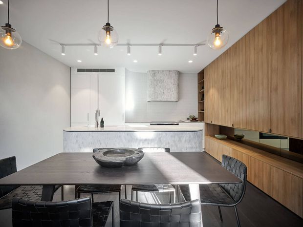 Contemporary open-plan kitchen and dining area with a long dark dining table and woven black chairs in the foreground. The kitchen features a pale stone island and benchtop, white full-height cabinetry, a textured pale splashback or rangehood wall, and extensive warm timber cabinetry along the right wall. Globe pendant lights and track lighting illuminate the clean, refined space.