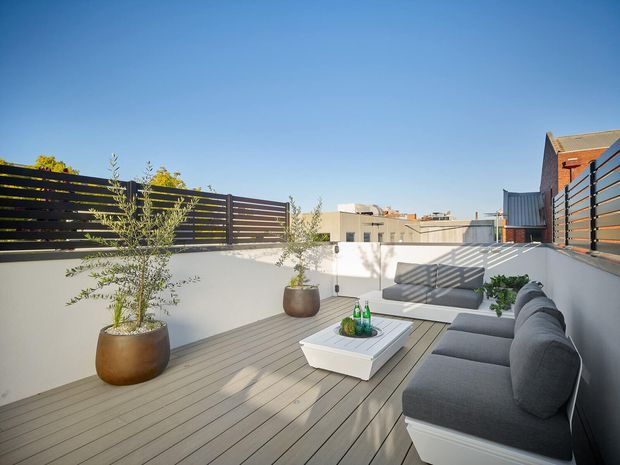 A sunny elevated outdoor terrace with grey composite or timber-look decking, white rendered parapet walls, dark horizontal privacy screening, and a built-in grey upholstered outdoor bench. Large potted olive-like trees, a low white coffee table, and simple refreshments create a contemporary rooftop entertaining space. Urban buildings are visible beyond the terrace.