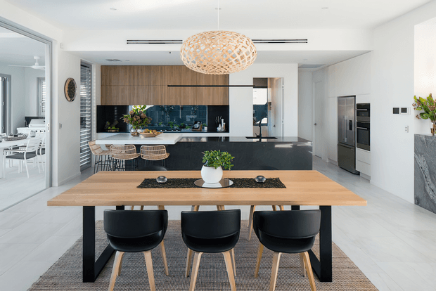 Bright, open-plan contemporary kitchen and dining area with a large light timber dining table, black dining chairs, a black kitchen island, timber cabinetry, dark splashback, integrated appliances and a sculptural woven pendant light. White walls, pale flooring and abundant glazing create an airy, refined interior.