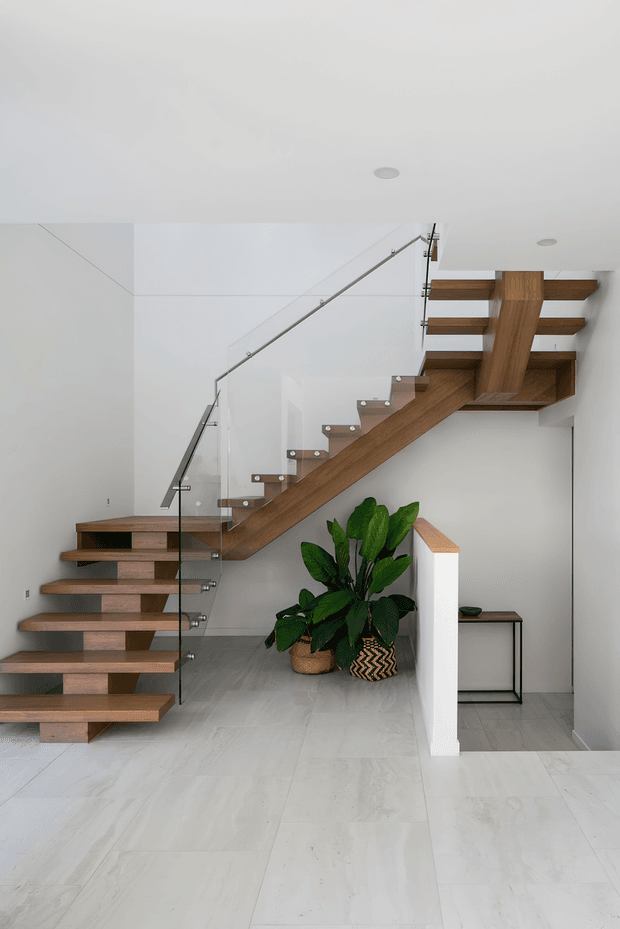 Bright minimalist interior stairway featuring an open-riser timber staircase with a prominent central timber spine and clear glass balustrades secured with metal fittings. The stairs connect to an upper landing, with white walls, recessed ceiling lights, pale large-format flooring, and indoor plants beneath the stair. A small console table is visible in the adjacent area.