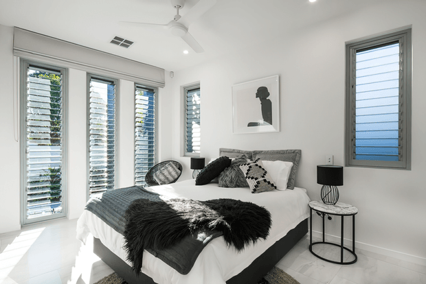Bright contemporary bedroom with a neatly made bed, layered white and charcoal bedding, textured dark throw, patterned cushions, black wire-frame bedside tables and lamps, and monochrome wall art. White walls and flooring are complemented by multiple grey-framed louvred windows that provide strong natural light. A ceiling fan and recessed lighting complete the room.