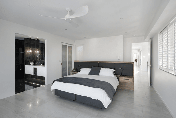Bright, contemporary bedroom with a large upholstered dark grey bed, white bedding, integrated dark bedside cabinetry, and a light timber bedside drawer. The room has white walls, pale tiled flooring, a sculptural white ceiling fan, plantation shutters, and a partially visible glossy black-tiled ensuite or bathroom through the opening on the left.
