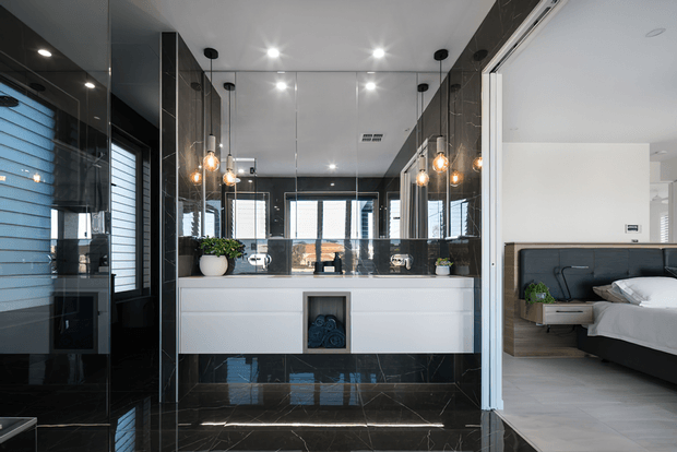Contemporary ensuite bathroom with a floating white double vanity, large mirrored wall, glossy black marble-look wall and floor surfaces, and warm globe pendant lights. A bedroom with a dark upholstered bed is visible directly through the open connection on the right. Black-framed windows and reflective surfaces create a dramatic, high-contrast finish.