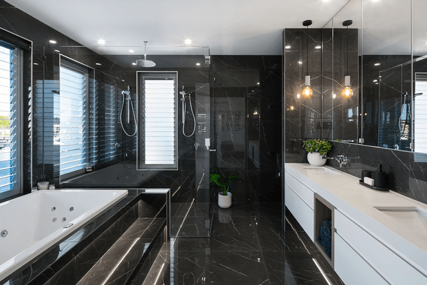 A luxurious contemporary bathroom with glossy black marble-look wall and floor tiles, a freestanding spa bath set into a raised tiled platform, twin wall-mounted showers, louvred windows, and a long white double vanity. Mirrored cabinetry and pendant lights add brightness and depth, while potted greenery provides a small natural accent.