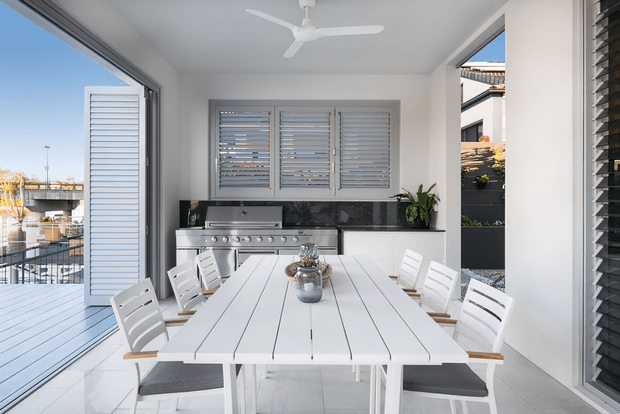 Bright, airy outdoor entertaining room with a large white dining table and chairs, built-in stainless-steel barbecue, louvred windows and shutters, white tiled flooring, ceiling fan, and extensive glazed openings connecting to a deck and waterfront-facing exterior.