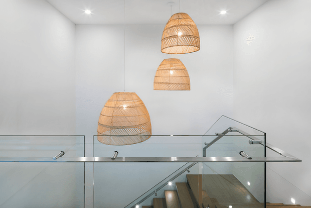 Bright, minimalist stair landing with clear glass balustrades, stainless-steel handrail details, and a stairwell descending below. Three woven natural-fibre pendant lights hang at different heights against white walls and ceiling, creating a warm organic contrast to the crisp contemporary architecture.