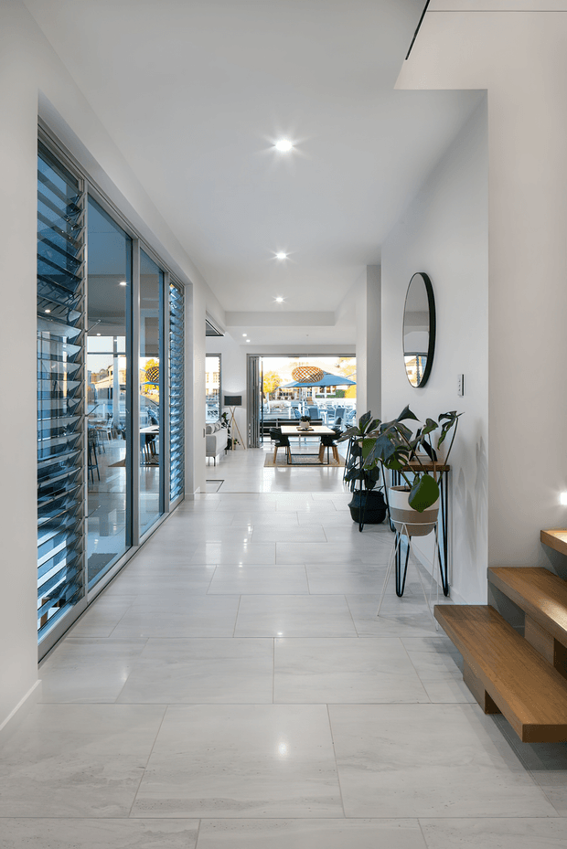 Bright contemporary residential hallway and entry circulation space with large floor-to-ceiling glazed louvred doors along the left, white walls and ceiling, polished pale stone-look tiled flooring, recessed downlights, a round wall mirror, indoor plants, and floating timber stairs on the right. A furnished dining and living area is visible at the far end.