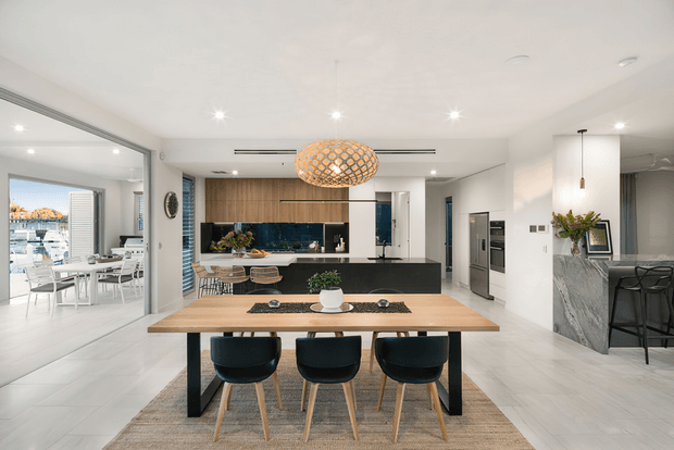 Bright, open-plan contemporary kitchen and dining area with a timber dining table, three dark upholstered chairs, a large island with dark cabinetry, white walls, pale stone-look flooring, and integrated appliances. A sculptural woven pendant hangs above the dining table, while a marble-look bar or benchtop with bar stools is visible on the right. Full-height glazing opens toward an outdoor waterfront or marina setting.