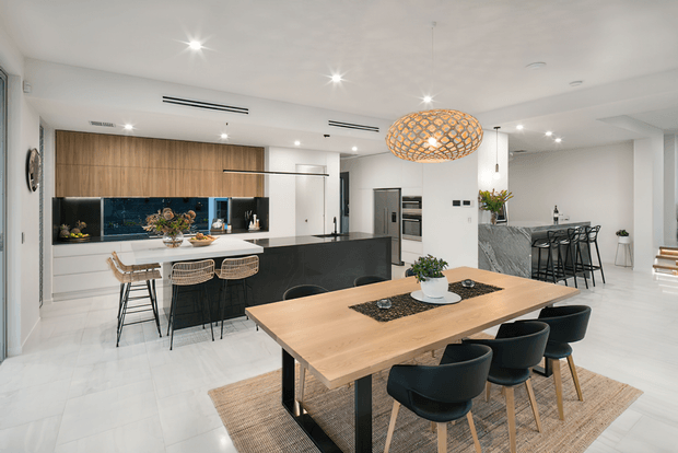 Bright open-plan kitchen and dining area with a large timber dining table, black upholstered dining chairs, a black kitchen island with bar seating, timber cabinetry, dark splashback, integrated appliances, and a sculptural woven pendant light. The space has white walls and pale flooring with a clean, contemporary fit-out.