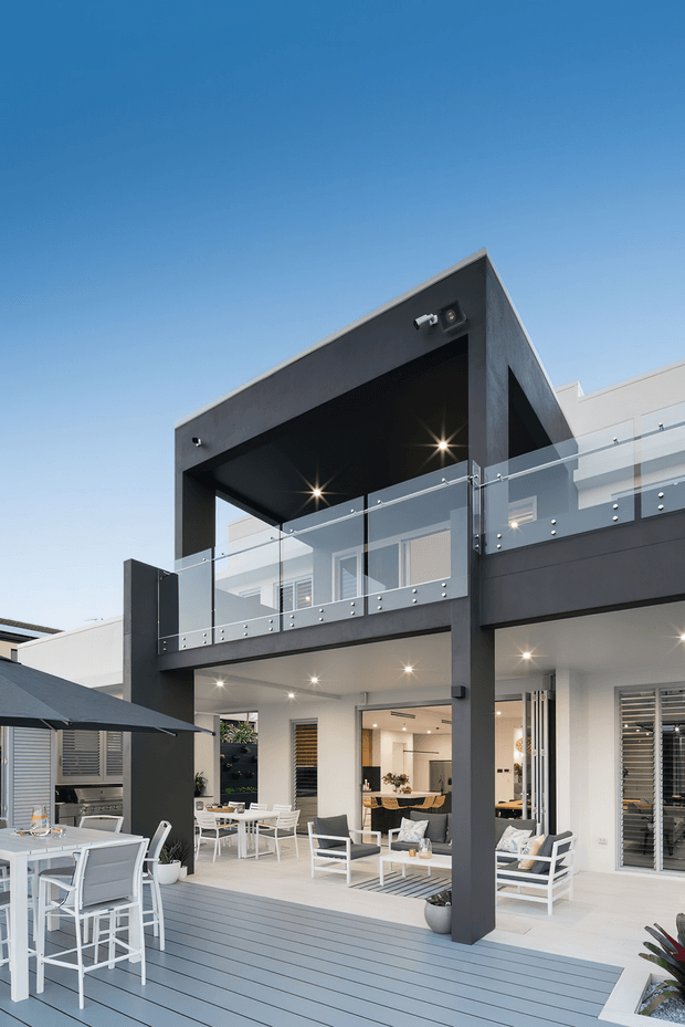 Contemporary multi-level residential exterior centered on a covered outdoor living and entertaining area. The facade combines dark charcoal structural framing with white walls, extensive glazing, and clear glass balcony balustrades. A spacious grey composite deck includes outdoor lounge seating and a bar-height dining setting, while the open upper-level terrace and recessed lighting emphasize the home's indoor-outdoor connection.