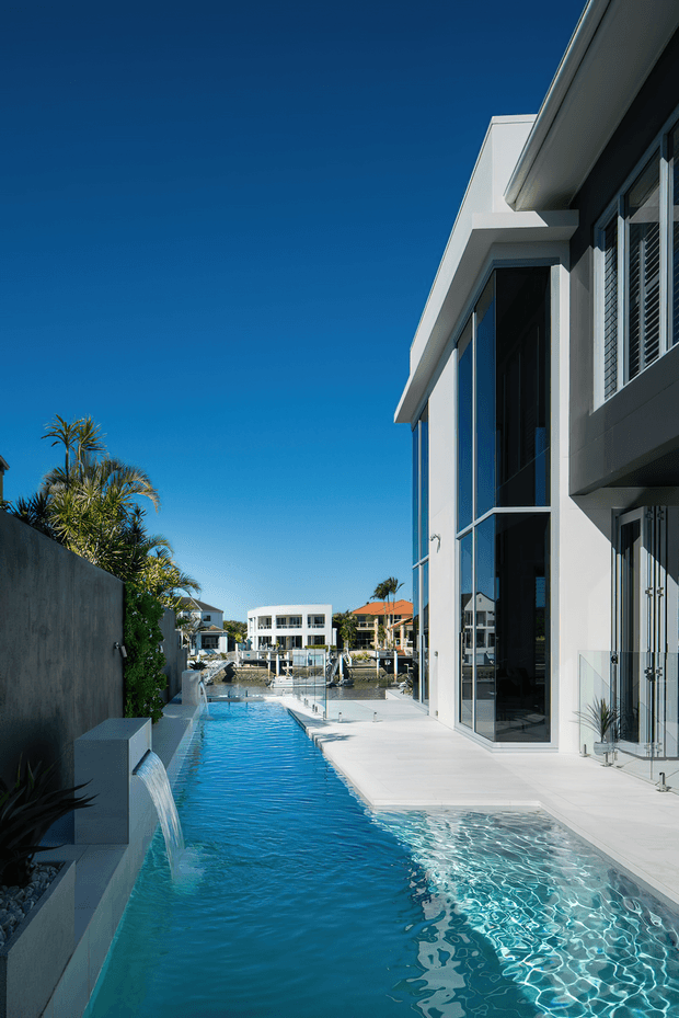 Exterior view of a contemporary multi-storey waterfront residence with a long narrow swimming pool running alongside the building. The house has crisp white rendered walls, extensive floor-to-ceiling glazing, dark window frames, projecting flat roof forms, and a pale paved pool deck. A water feature feeds into the pool, with neighboring waterfront homes visible beyond.