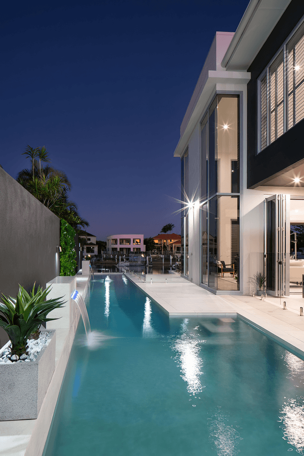 Dusk exterior view of a contemporary multi-level residence beside a long narrow swimming pool. The property features white rendered walls, extensive floor-to-ceiling glazing, dark screening and a projecting roofline. A raised planter wall with tropical planting borders the pool, which includes a small water feature and illuminated water surface. Interior spaces are visible through the open glazed doors, with waterfront residences in the background.