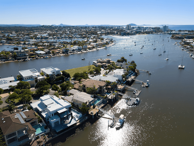 Aerial view of a waterfront suburban neighbourhood surrounding a broad river or estuary, with numerous houses, private docks, moored boats, and a denser urban skyline in the distance. The image is a bright daytime exterior landscape photograph.