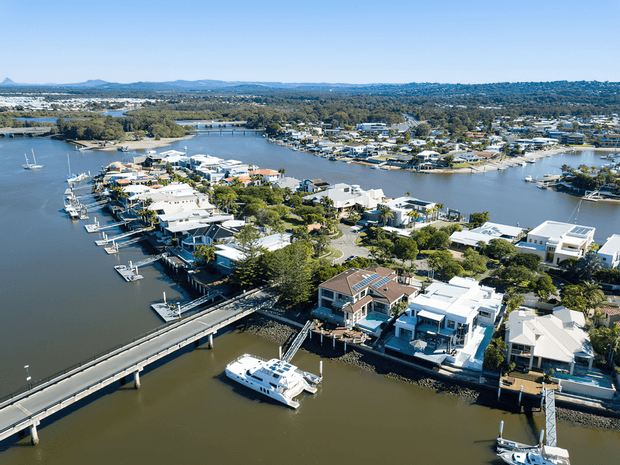 Aerial view of a low-density waterfront residential neighbourhood built along a broad river or estuary, with numerous homes, private jetties, moored boats, landscaped gardens, and a road bridge crossing the water. The scene has a sunny coastal-suburban character with distant hills and extensive shoreline development.