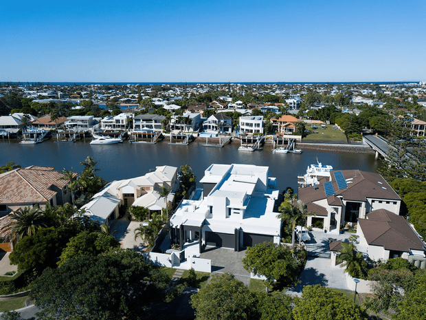 Aerial view of a suburban waterfront residential neighbourhood, showing several large detached homes, private docks, boats, landscaped yards, and a calm canal leading toward a distant coastal horizon. The foreground includes a prominent white contemporary flat-roof house and neighbouring homes with pitched roofs.