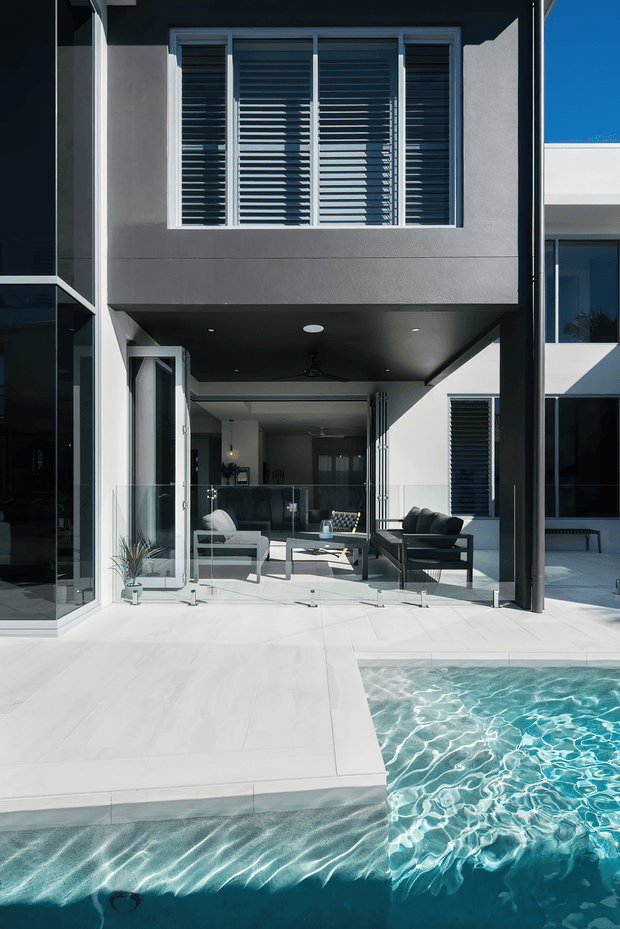 Bright contemporary multi-storey residence viewed from the pool courtyard, with white tiled paving, a turquoise swimming pool, expansive glazing, a covered outdoor living area, dark structural columns, and upper-level windows fitted with horizontal louvres.