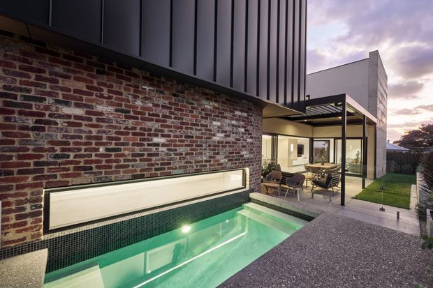 A contemporary residential courtyard viewed at dusk, featuring a narrow illuminated swimming pool beside a textured exposed-brick wall, dark vertical metal cladding above, and a glazed outdoor dining pavilion. The paved courtyard extends toward a small lawn and timber fence, with warm interior lighting visible through the glazing.