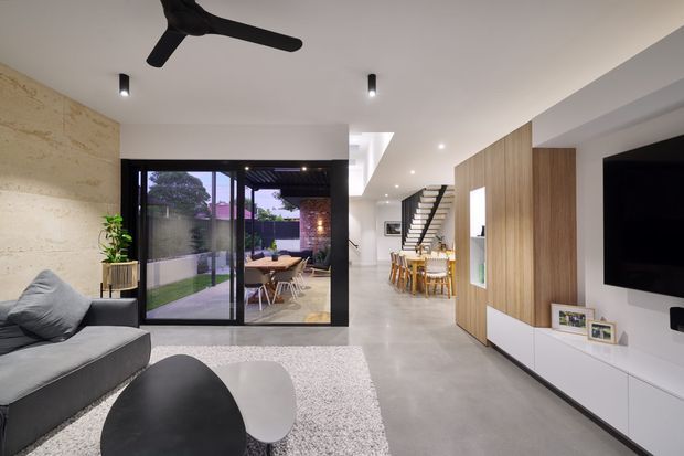 Contemporary open-plan living room with polished concrete flooring, a neutral grey sofa, black ceiling fan, large sliding glass doors opening to a covered outdoor dining area, and a timber feature wall incorporating white media cabinetry and a television. The dining area and staircase are visible beyond the living space.